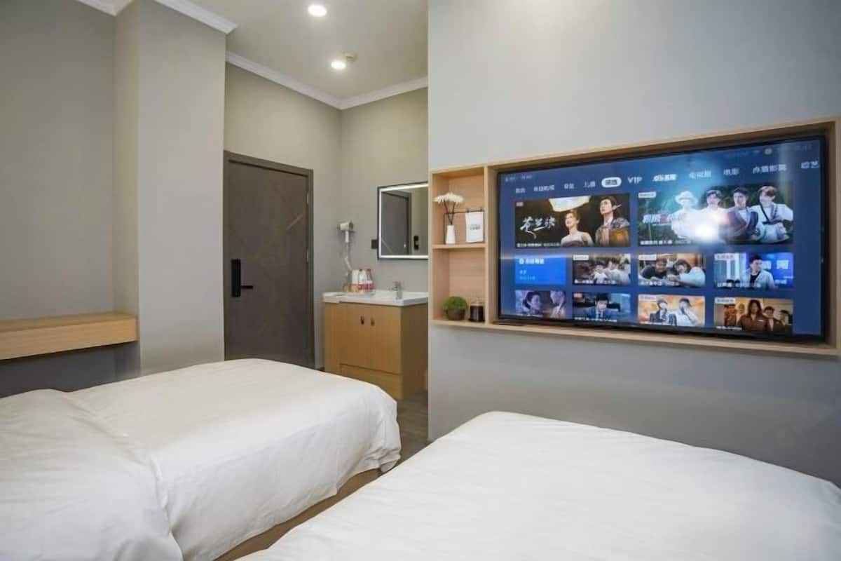 Quarto de hotel com duas camas, uma televisão fixada na parede exibindo diversos programas, um pequeno estante com artigos de higiene e uma porta que dá para outro cômodo.