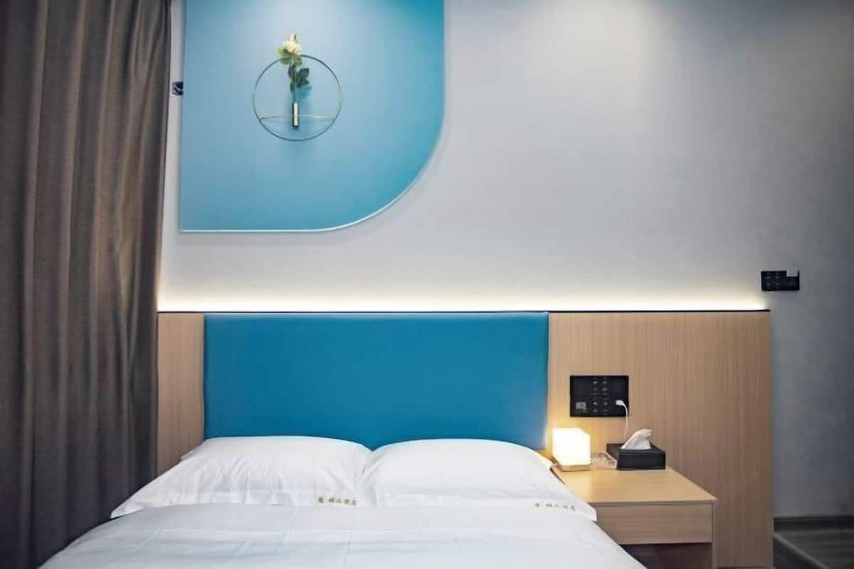 Quarto de hotel com uma cama, uma mesinha de cabeceira com abajur, um painel de controle fixado na parede e um vaso de flores na parede.