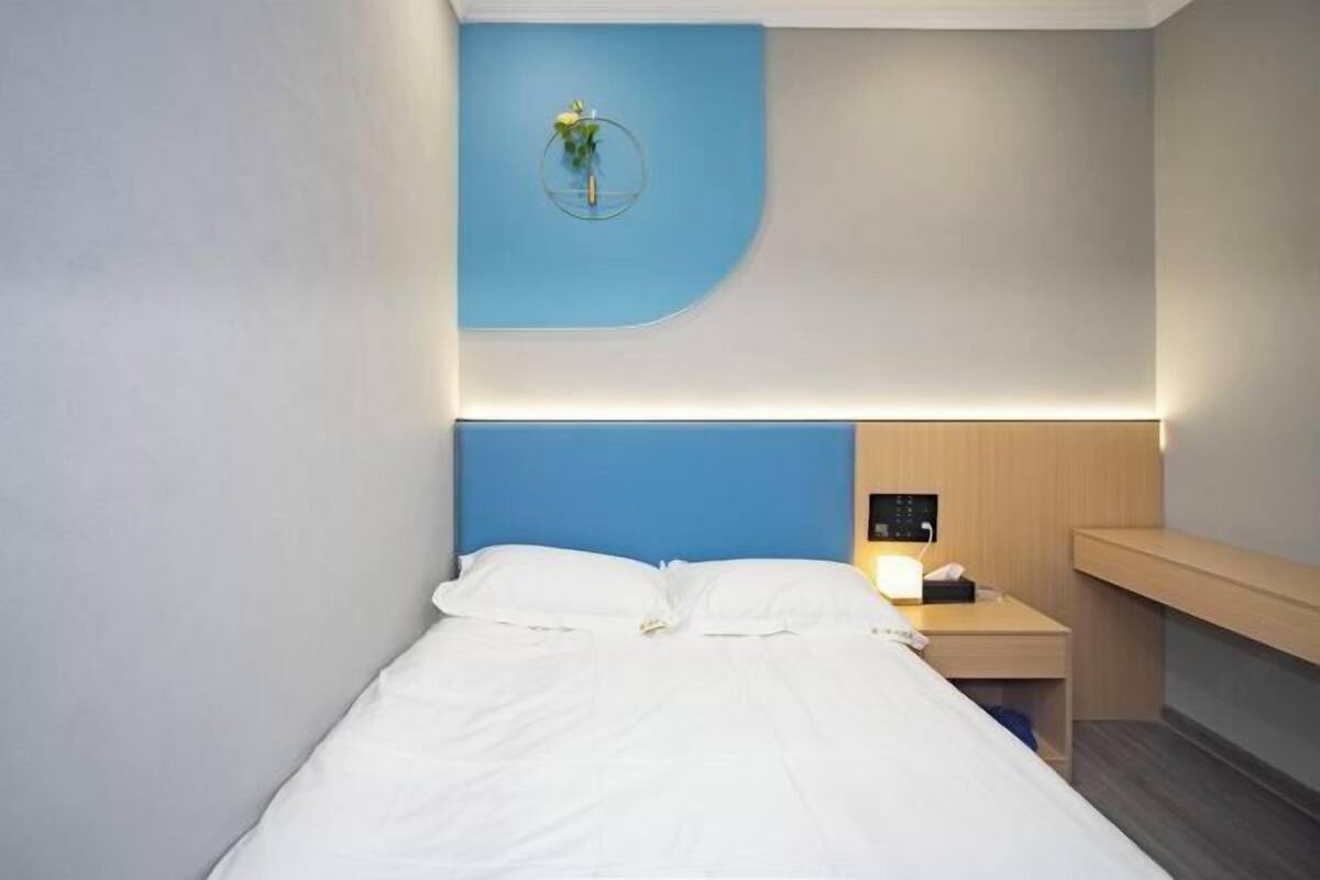 Quarto de hotel moderno com cama, mesa de cabeceira com luminária, pequena escrivaninha e luminária na parede.