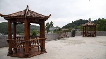 Two wooden gazebos with benches, one with a tiled roof and the other with a curved roof, on a rooftop with a view of a hilly landscape.