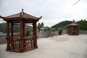 Two wooden gazebos with benches, one with a tiled roof and the other with a curved roof, on a rooftop with a view of a hilly landscape.