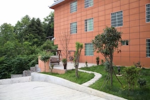 A building with a red facade, multiple windows, and a landscaped garden area with trees and a pathway. - Junlong Holiday Hotel (Dafang)
