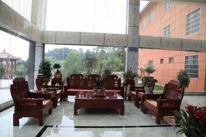 A lobby with a high ceiling, a central coffee table surrounded by chairs, and a view of a building and trees outside. - Junlong Holiday Hotel (Dafang)