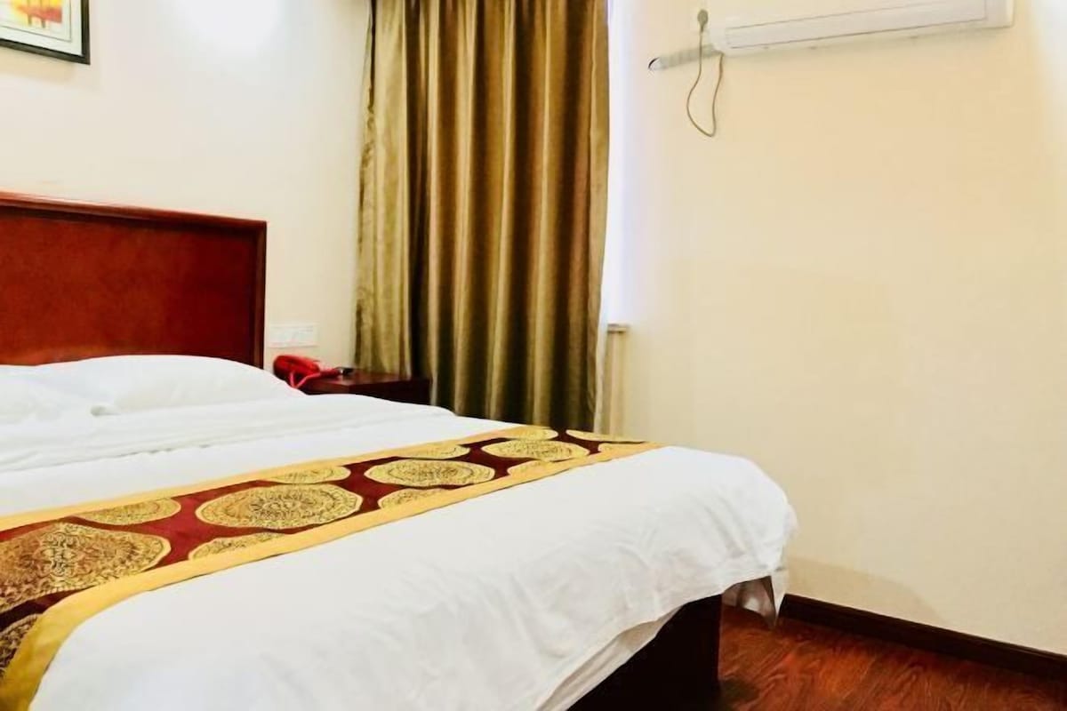 A hotel room with a bed, a wooden headboard, a window with curtains, and a wall-mounted air conditioning unit.