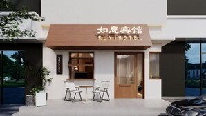 Ingresso moderno di un hotel con un cartello che recita 'RUYIHOTEL'. Due sedie e un tavolo sono posti all'esterno, e un'auto parcheggiata di fronte.