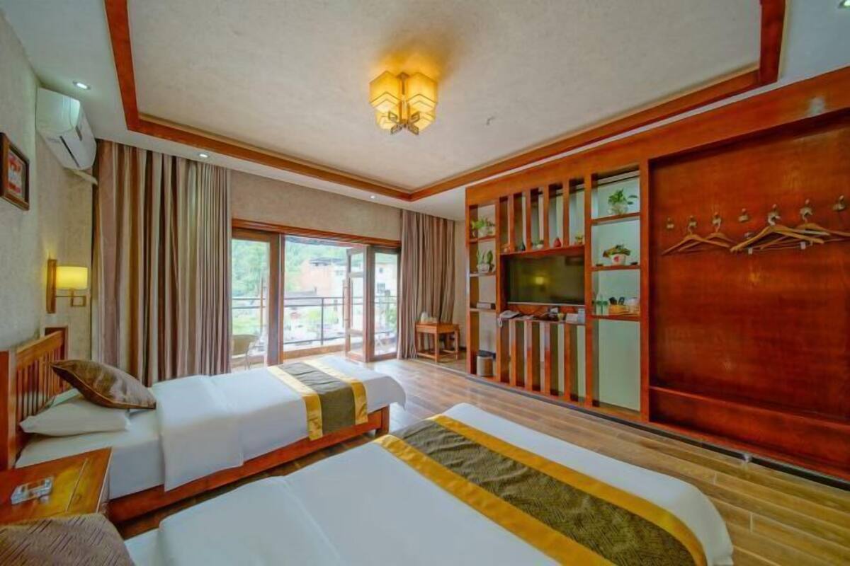 A hotel room with two beds, a television, a wooden cabinet with shelves, and a large window.