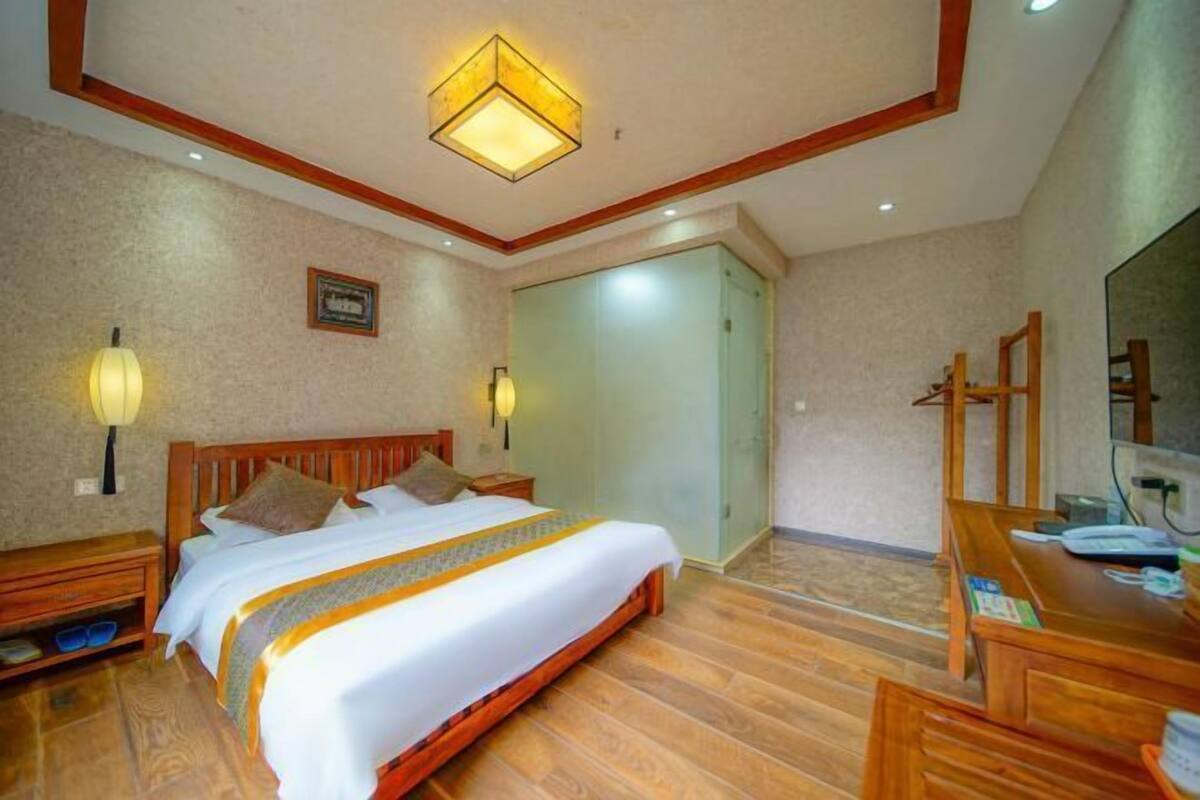 A hotel room with a wooden bed, bedside tables, a desk with a chair, a mirror, and a ceiling light fixture.
