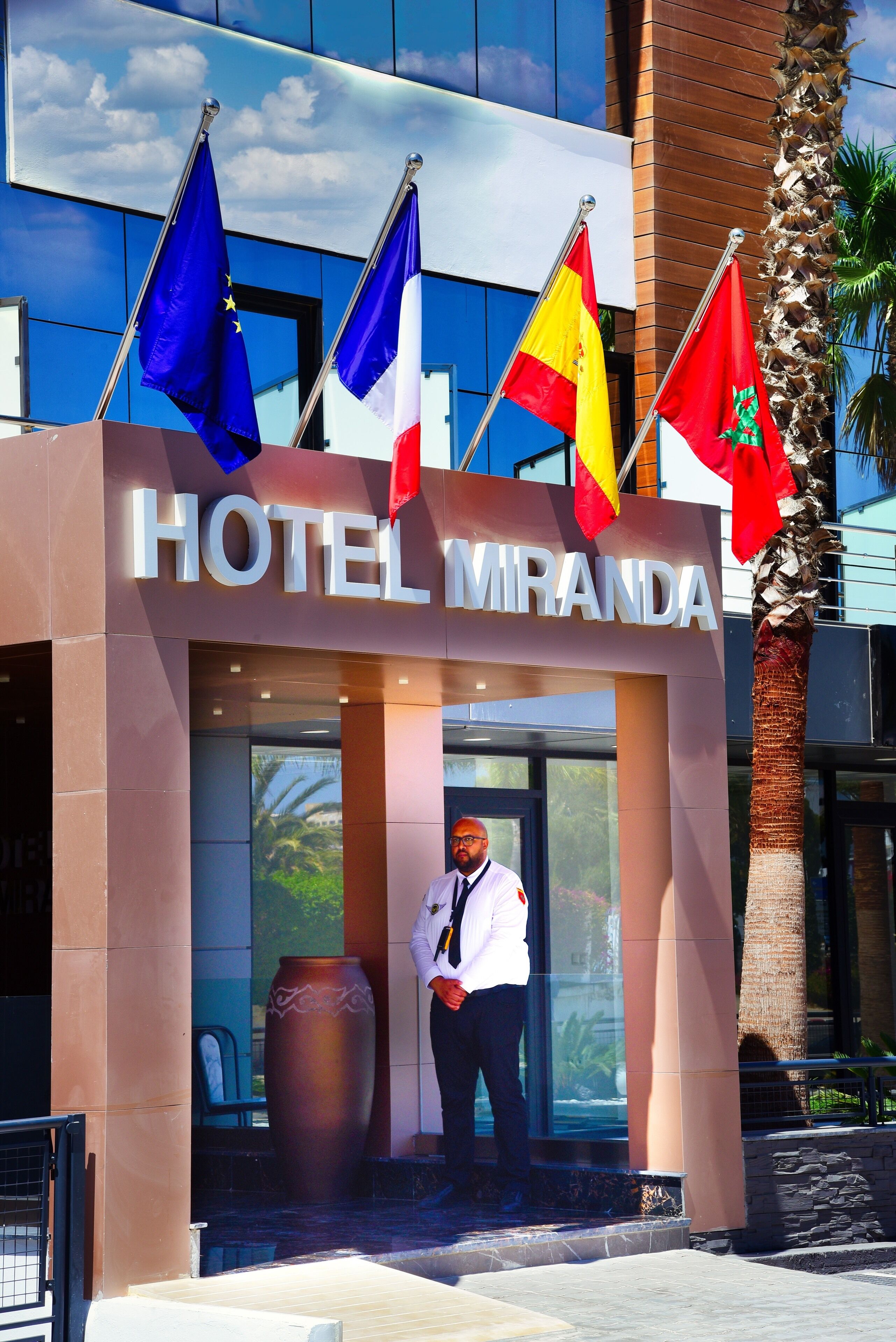 Entrada de un hotel con banderas, un cartel que dice "HOTEL MIRANDA" y un guardia de seguridad.