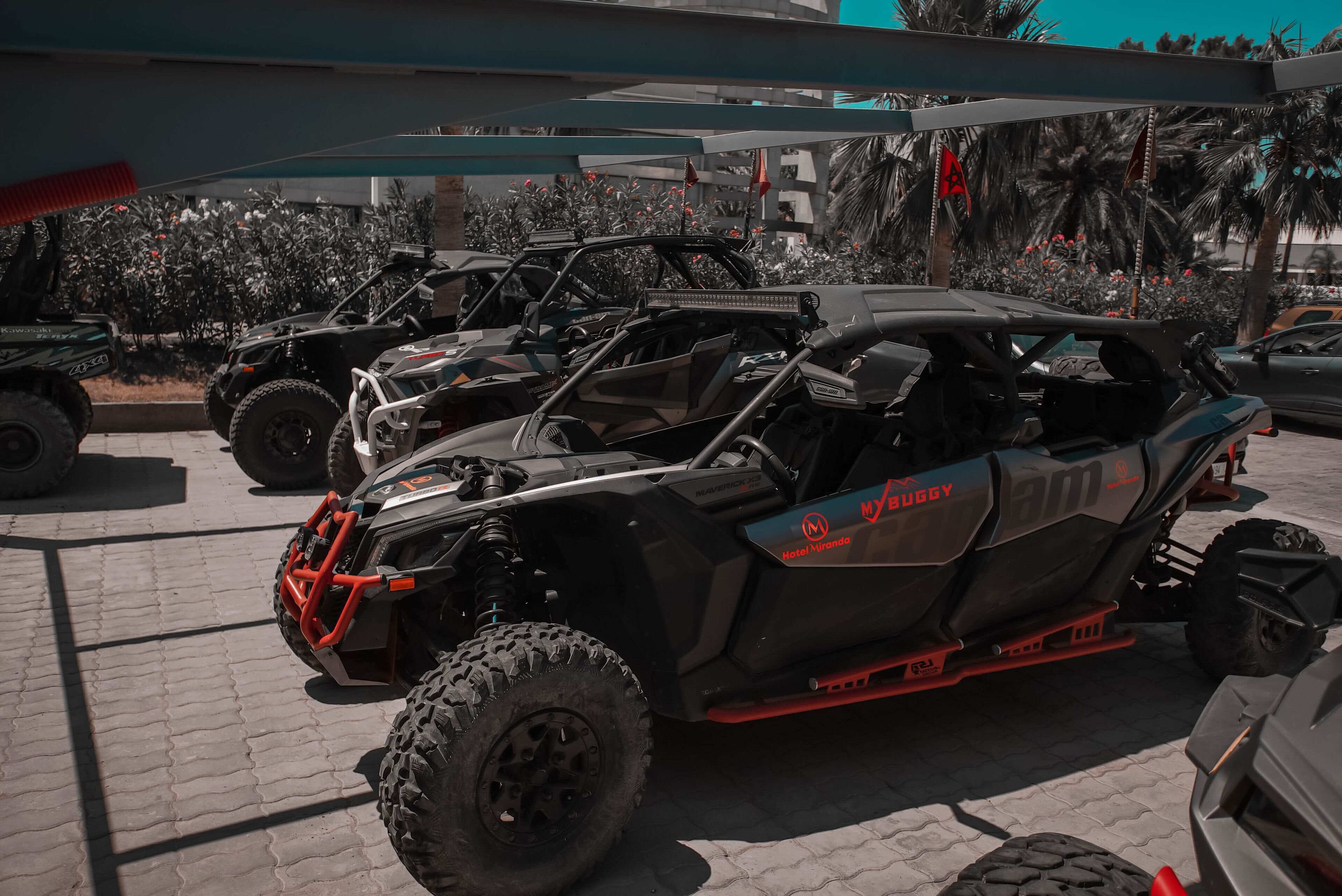 A black and red off-road vehicle with "MYUGGY" branding parked in a paved area with palm trees and a building in the background.