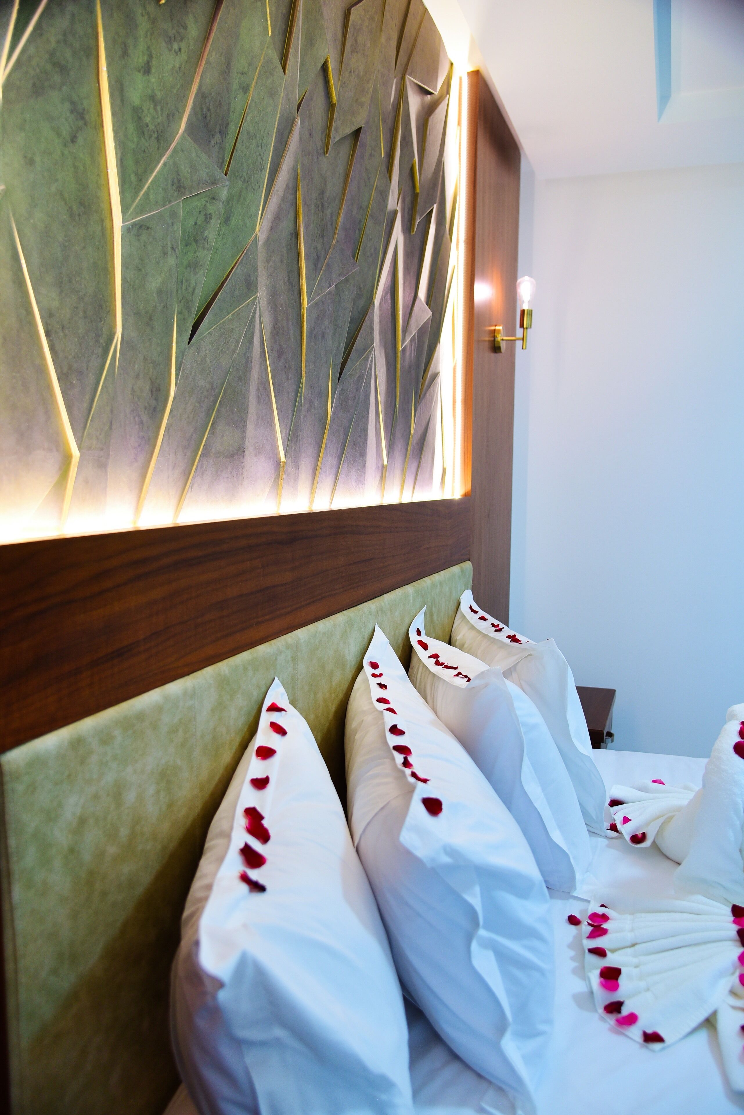 A hotel room with a modern headboard, neatly made beds with white linens and rose petals, and a wall-mounted light fixture.