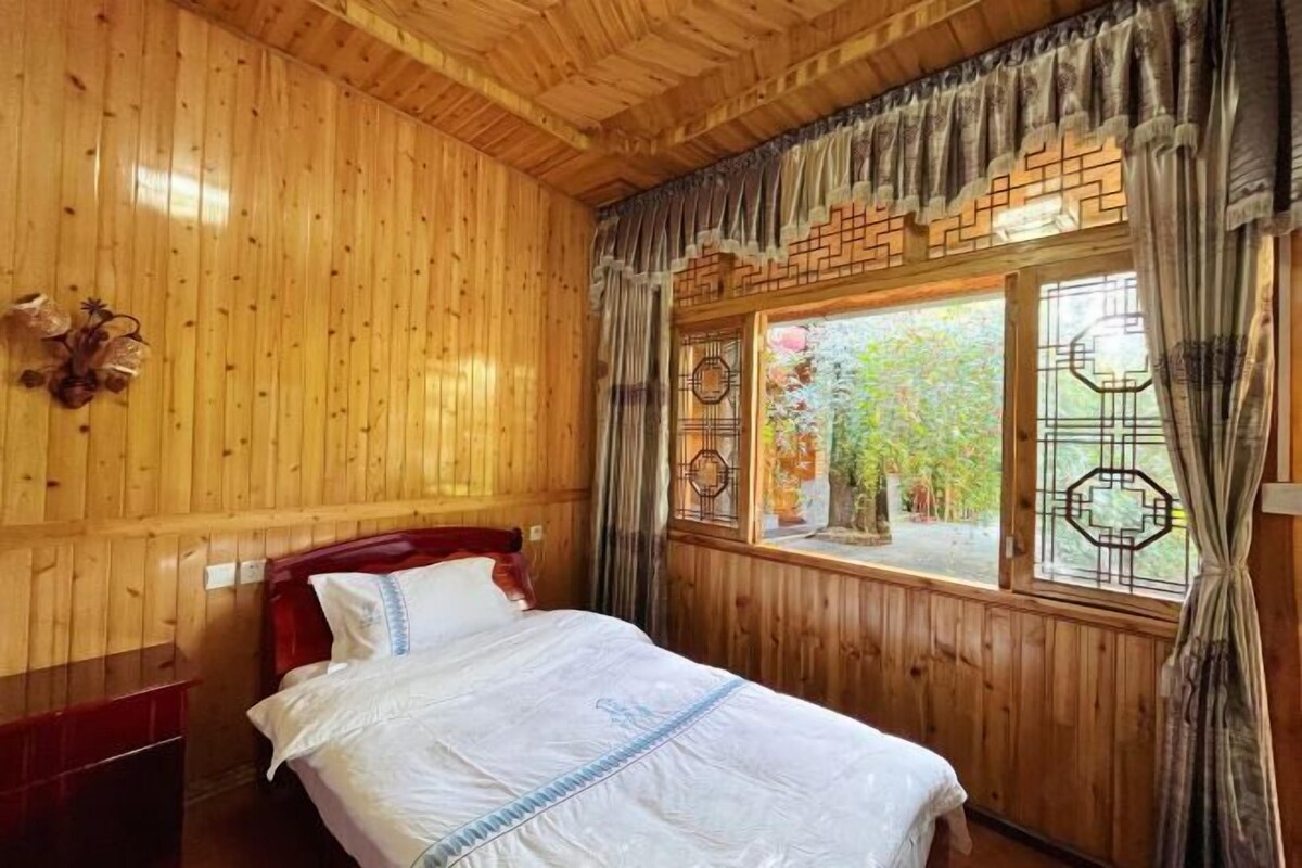 A wooden-paneled room with a bed, a nightstand, and a window with decorative curtains.