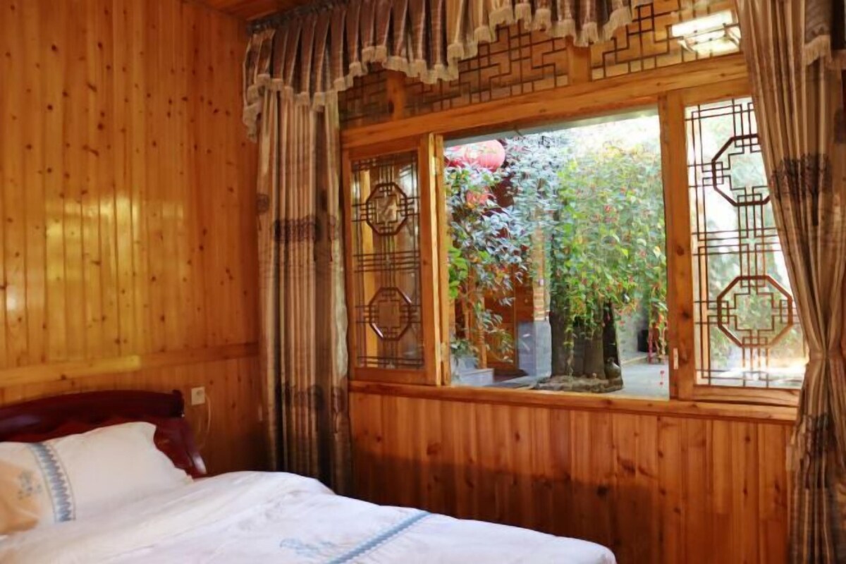 A wooden room with a bed, a window with decorative metal grilles, and a view of greenery outside.