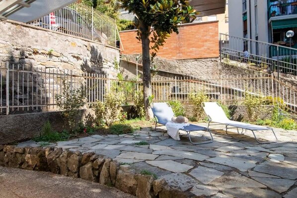 A stone patio with two lounge chairs and a tree, surrounded by a stone wall and metal fence.