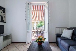 Un salon avec un canapé, une table basse ornée de fleurs, un téléviseur et une vue sur une terrasse extérieure dotée d’un auvent à rayures.