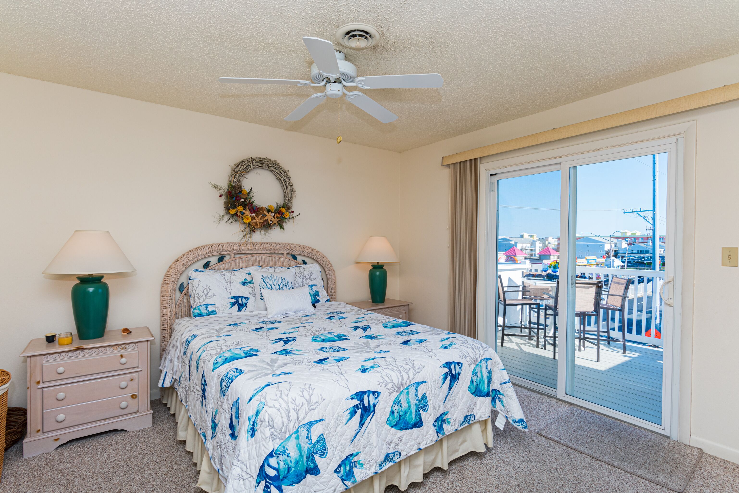 A bedroom with a bed, two nightstands, a ceiling fan, a window with a view of a balcony, and a wreath on the wall.