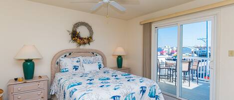 A bedroom with a bed, two nightstands, a ceiling fan, a window with a view of a balcony, and a wreath on the wall.
