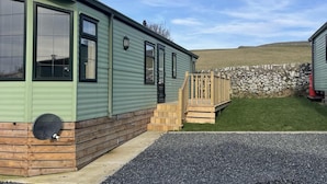 A green mobile home with a wooden deck and a stone wall in the background. - Immaculate 2-bed Static Caravan at Monrieth (Newton Stewart)
