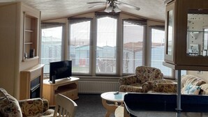 A living room with a TV, a ceiling fan, and a view of buildings through large windows. - Immaculate 2-bed Static Caravan at Monrieth (Newton Stewart)