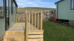 A wooden deck leading to a stone wall with a green building in the background. - Immaculate 2-bed Static Caravan at Monrieth (Newton Stewart)