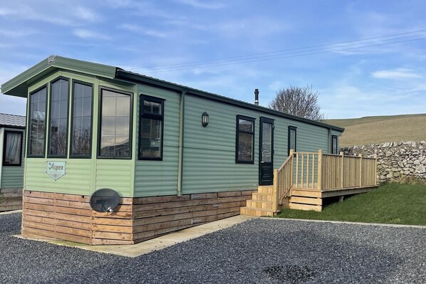 A single-story, light green mobile home with a wooden deck and a stone wall in the background. - Immaculate 2-bed Static Caravan at Monrieth (Newton Stewart)