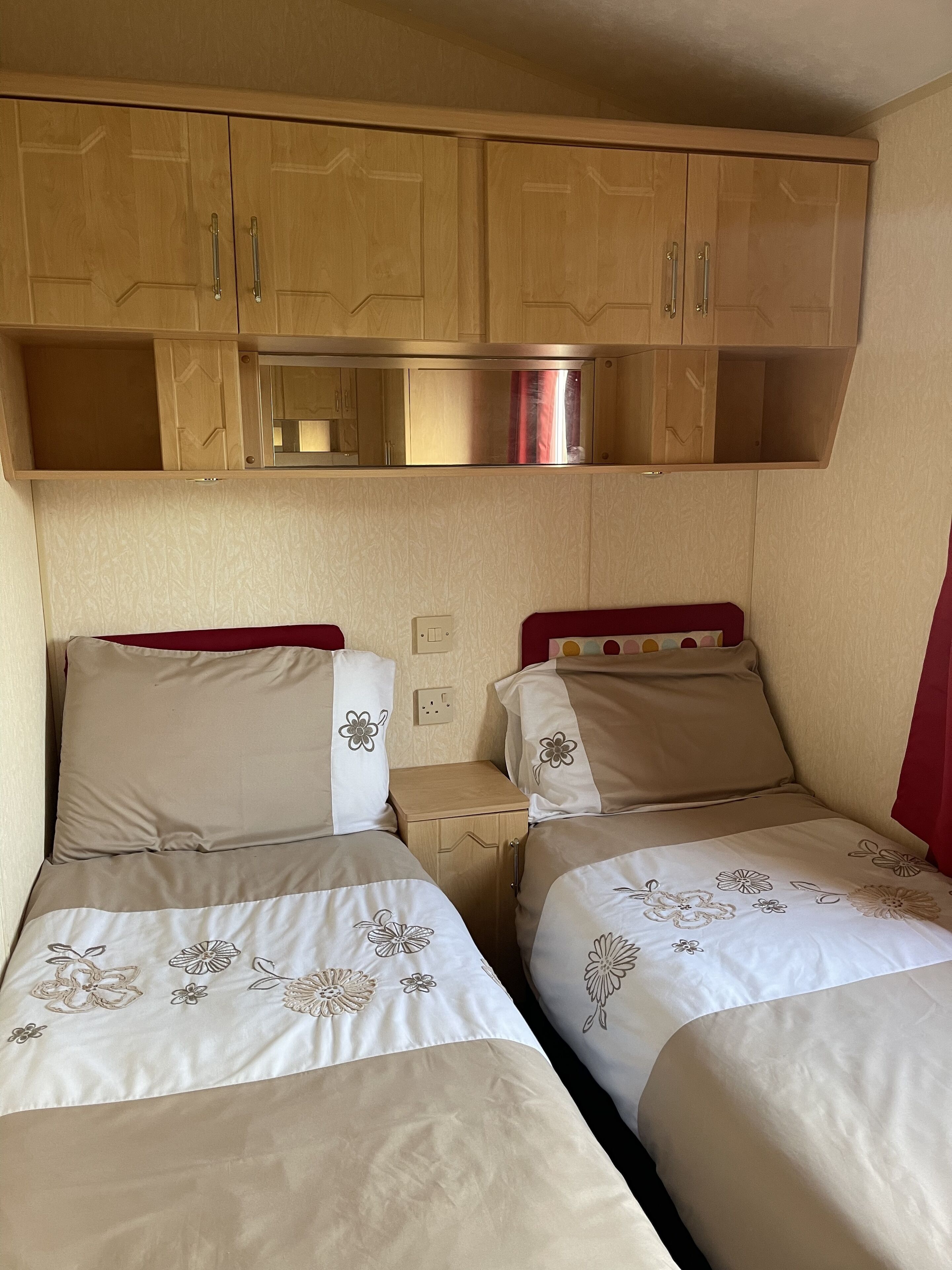 Habitación pequeña con dos camas, cada una con ropa de cama de motivos florales, armarios de madera arriba y una mesita de noche.