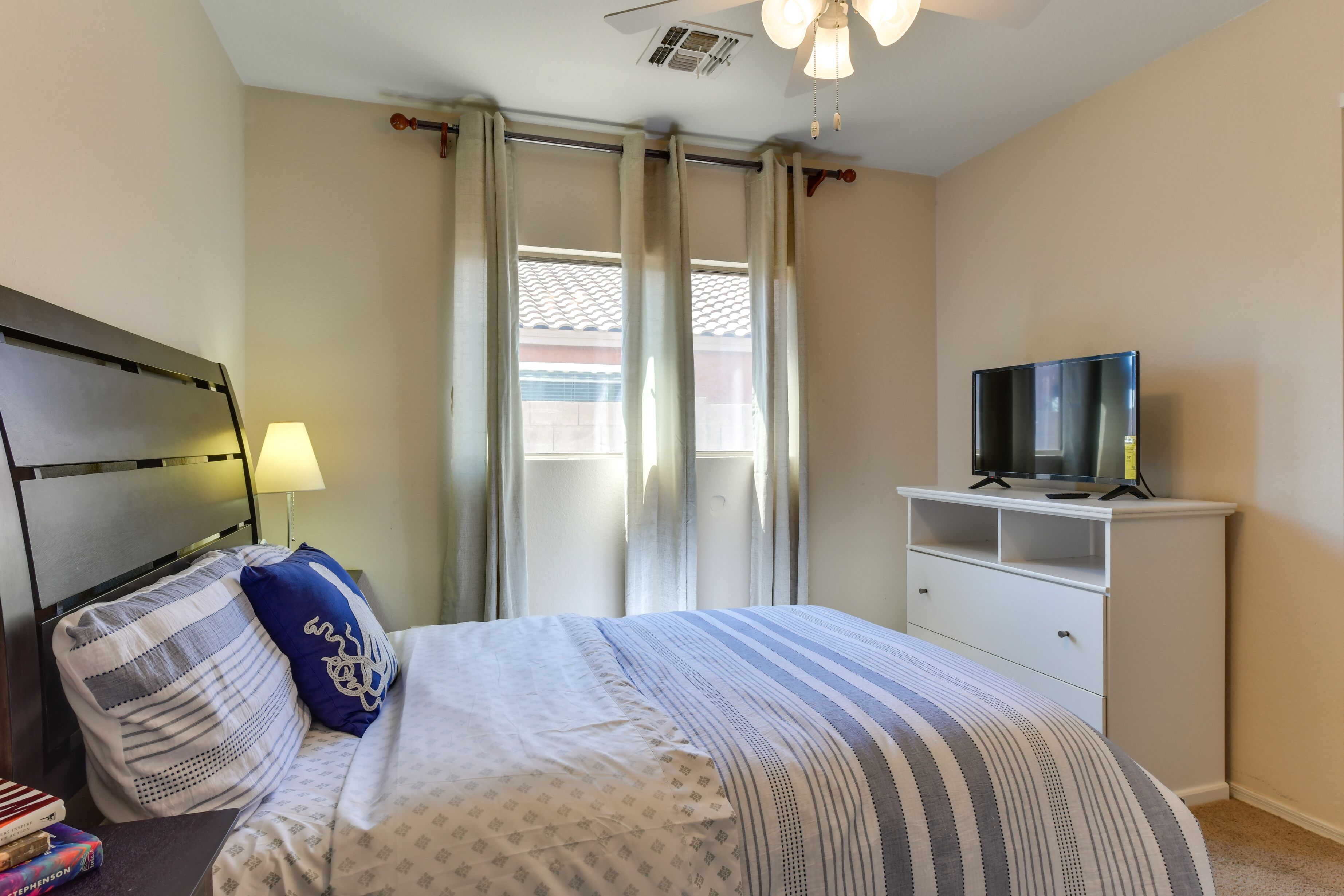 A bedroom with a bed, a TV on a white dresser, a lamp, and a window with curtains.
