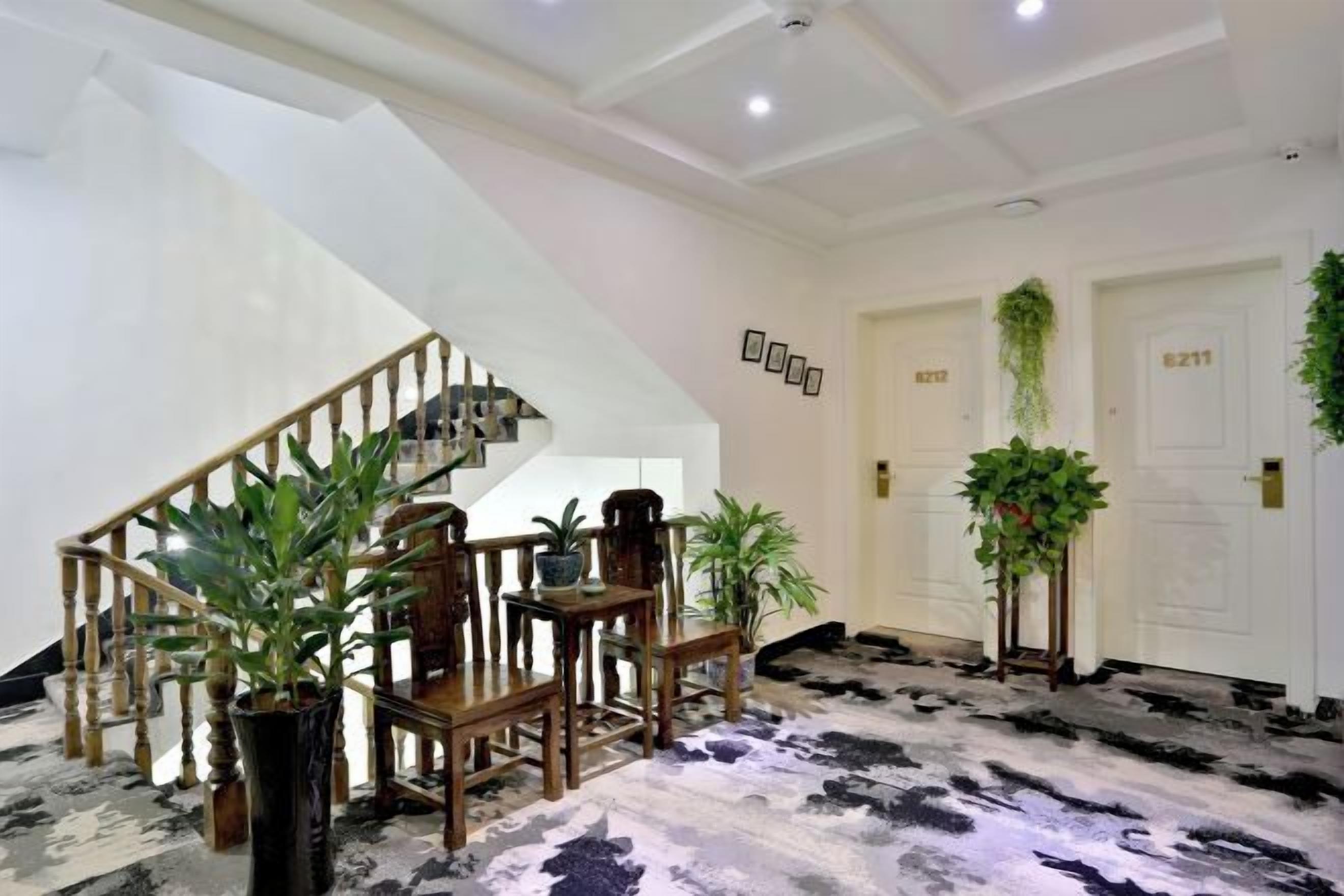 Vestíbulo de hotel con escalera, mobiliario de madera, plantas en macetas y alfombra estampada.