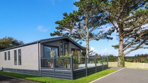 A modern mobile home with a balcony, a fenced garden area, and a large tree nearby. - Piran Lodge (Padstow)