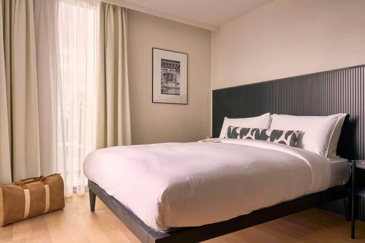 A neatly made bed with white linens and a dark headboard, a framed picture on the wall, and a wooden floor.