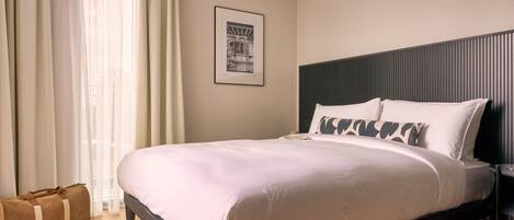 A neatly made bed with white linens and a dark headboard, a framed picture on the wall, and a wooden floor.