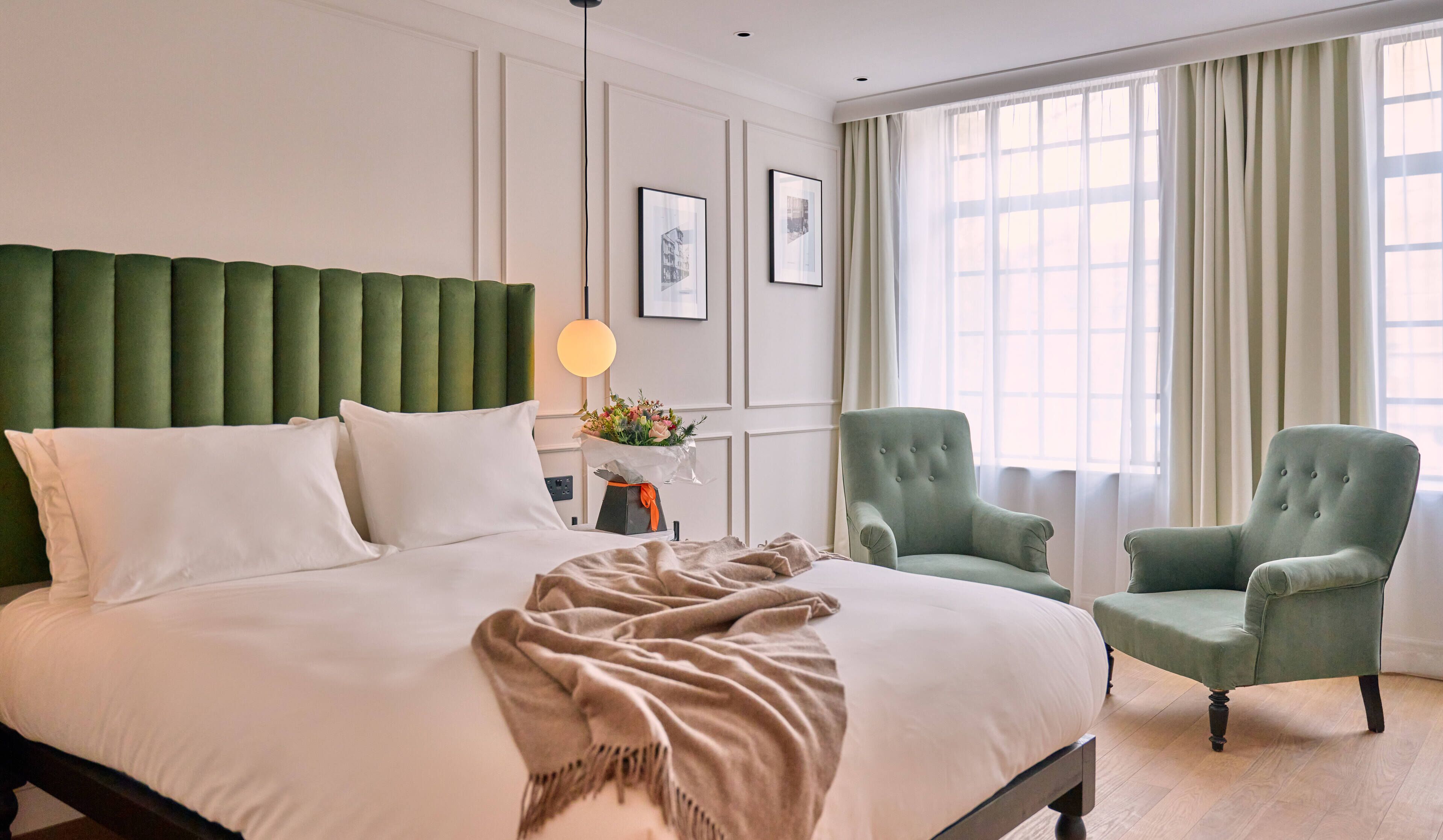 A neatly made bed with white linens, a green upholstered headboard, and a bedside table with a lamp and a bouquet of flowers. There are two green armchairs and a large window with sheer curtains.