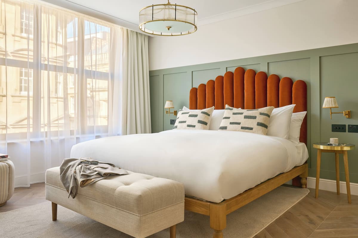 A neatly made bed with a wooden frame and a green headboard, a bedside table with a lamp, and a window with sheer curtains.