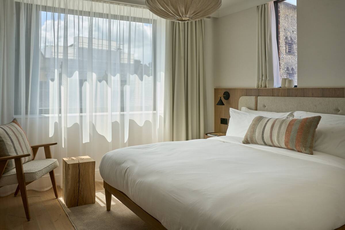 A neatly arranged bedroom with a large bed, a chair, a bedside table, and a window with sheer curtains.