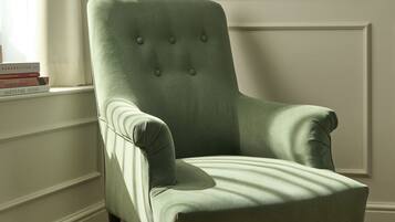 Un fauteuil vert, recouvert d’un tissu, doté d’un haut dossier et d’un capitonnage à boutons, placé dans un coin d’une pièce au plancher en bois et aux murs recouverts de panneaux blancs.