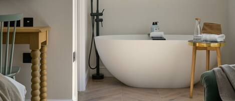 A modern bathroom with a freestanding bathtub, a wooden chair, a small round table with a towel, and a television mounted on the wall.