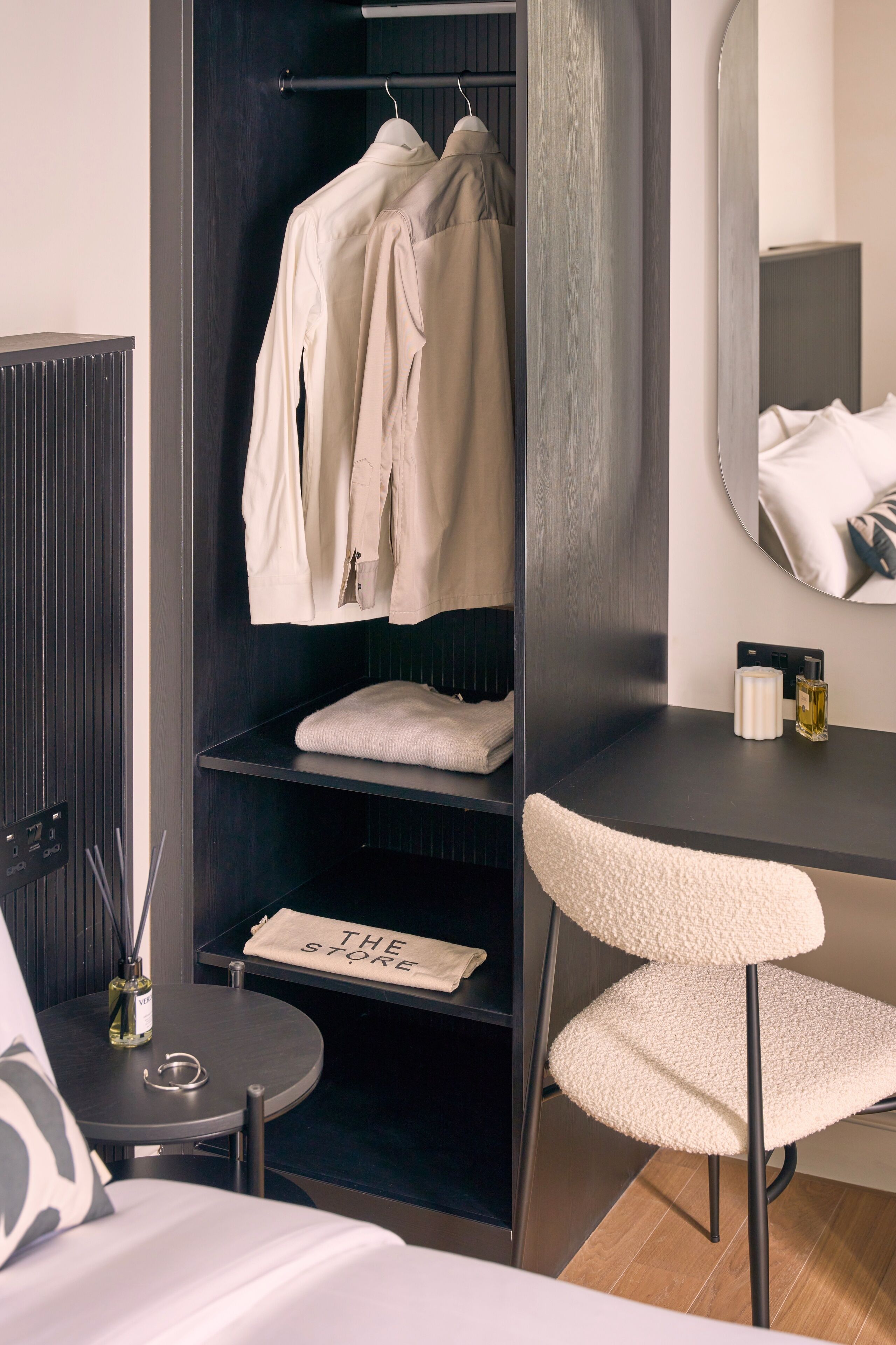 A hotel room with a dark wood wardrobe, a round mirror, a small table with a bottle and a candle, and a chair with a towel on it.