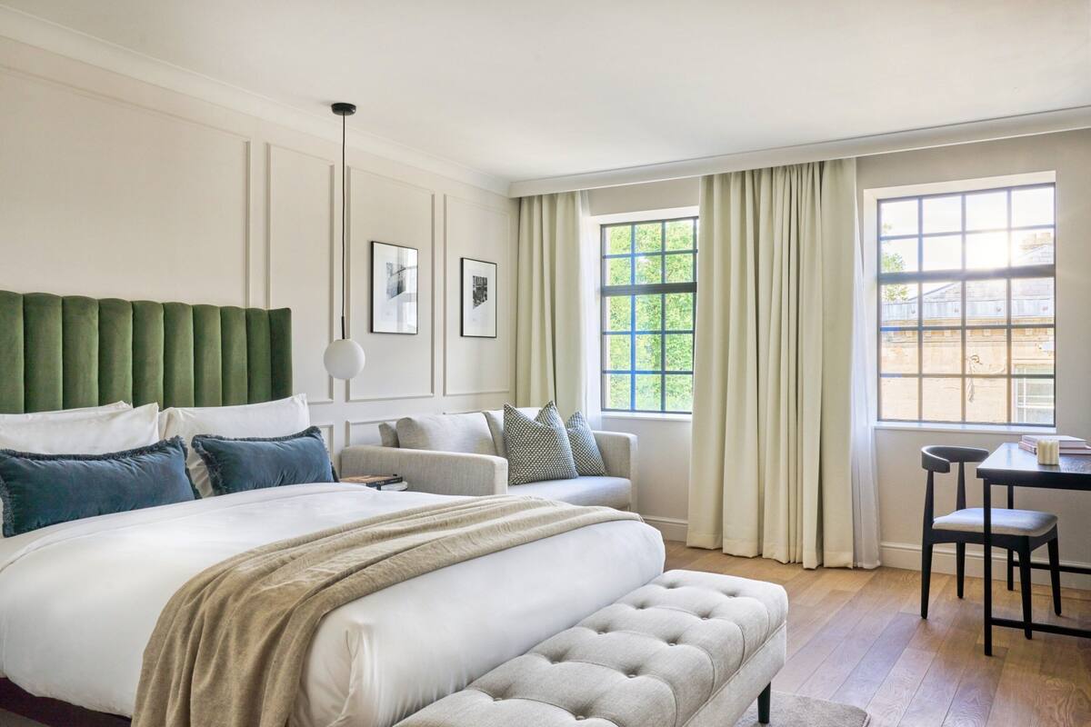 A hotel room with a large bed, a green upholstered headboard, a sofa, a table, and a window with curtains.