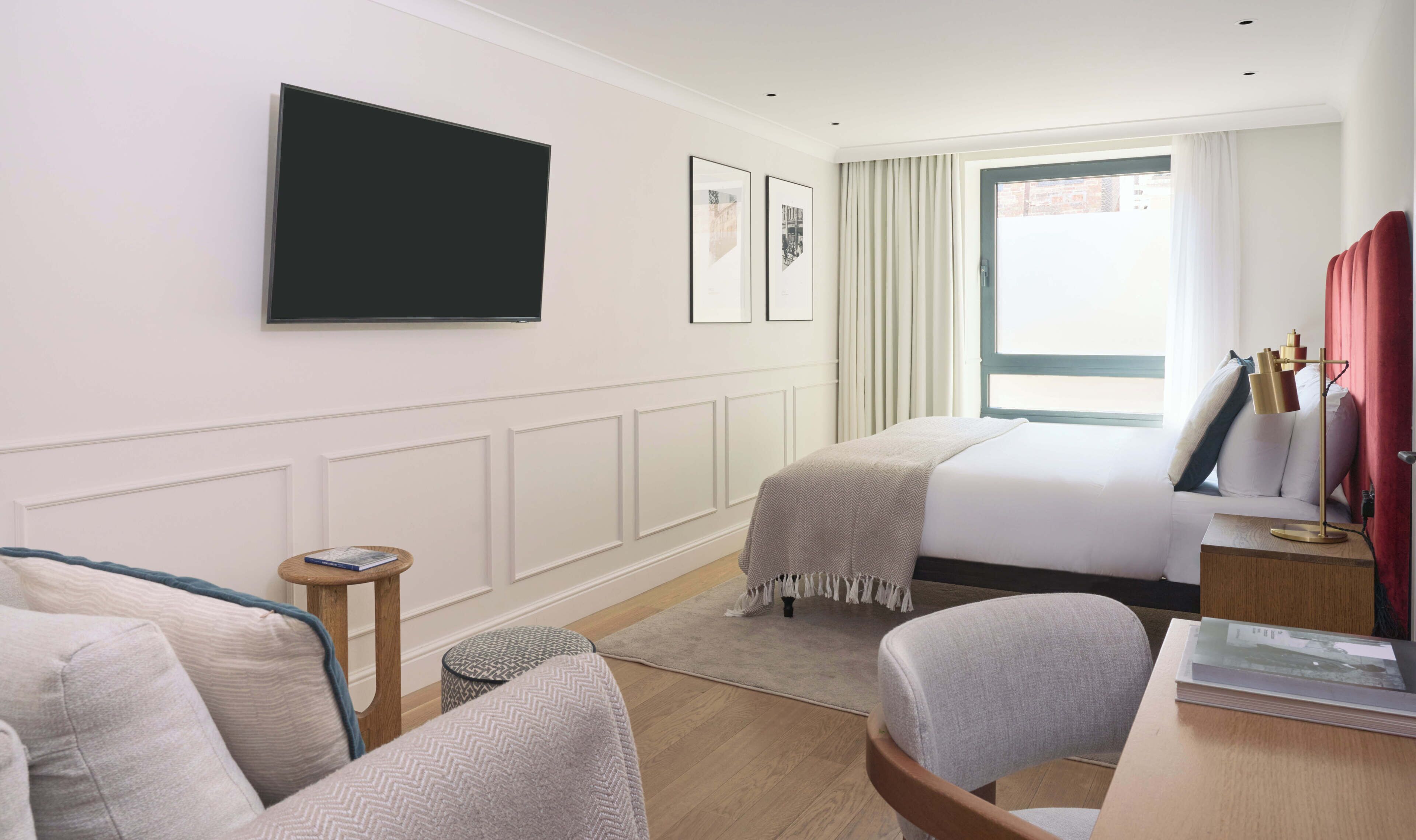 A hotel room with a bed, a desk with a lamp, a chair, a small round table, and a television mounted on the wall.