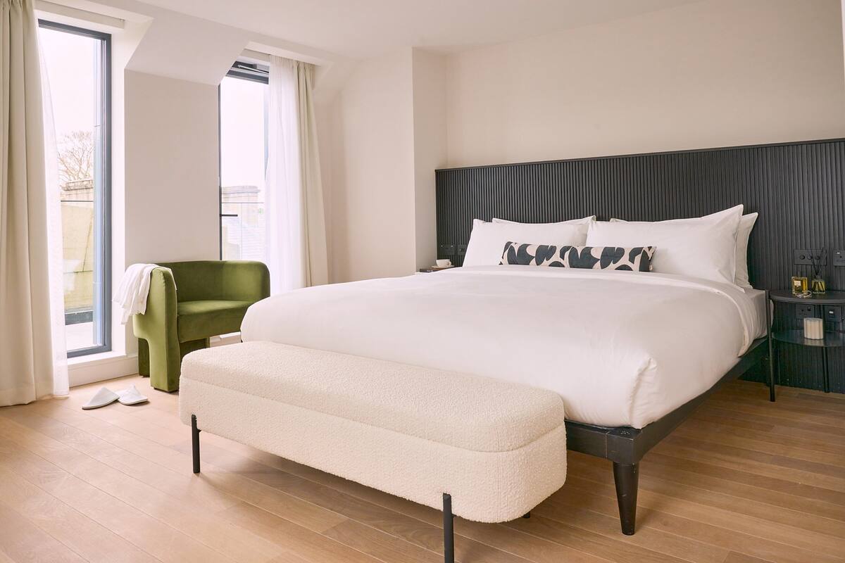 A modern hotel room with a large bed, a green armchair, and a view through a large window.