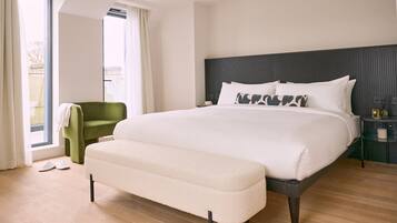 A modern hotel room with a large bed, a green armchair, and a view through a large window.