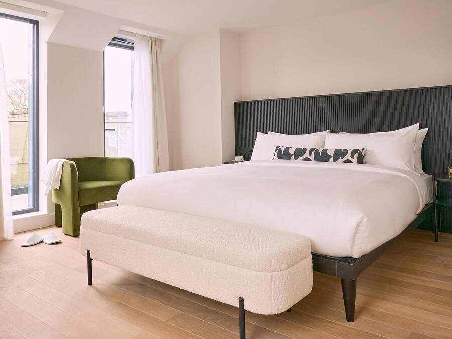 A modern hotel room with a large bed, a green armchair, and a view through a large window.