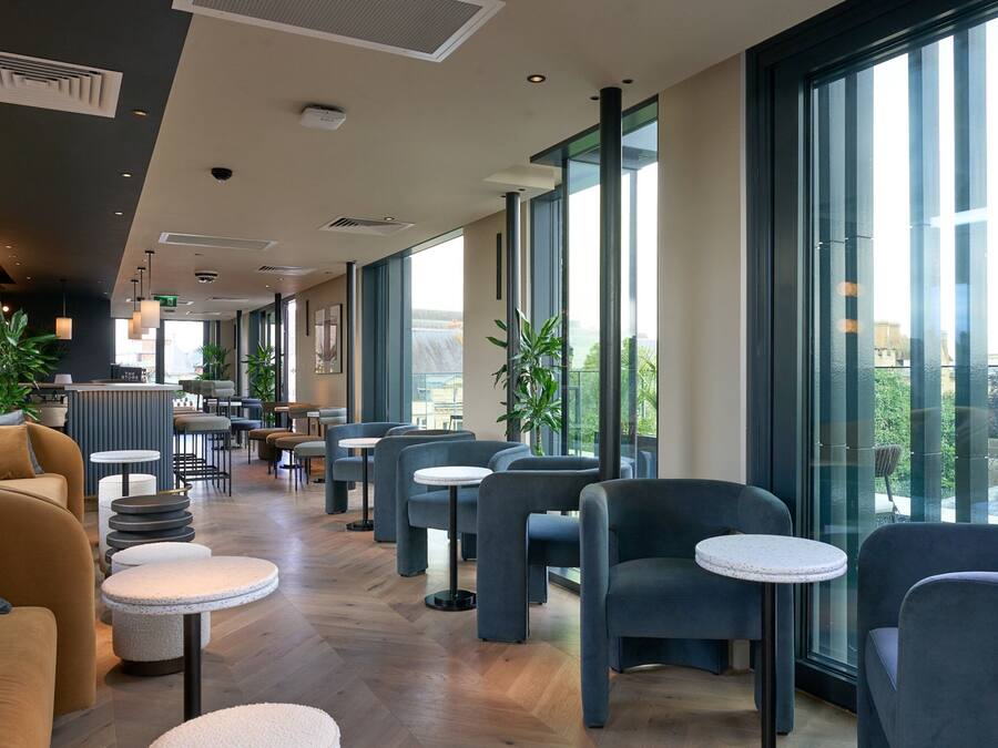 Una cafetería moderna con una barra larga, varias zonas de estar y grandes ventanales con vistas a la ciudad.