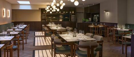 A restaurant with multiple dining tables and chairs, a bar area, and a kitchen in the background.