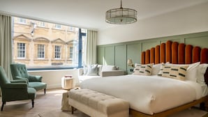 A bedroom with a large bed, a green armchair, a bench, and a view of a building through the window.