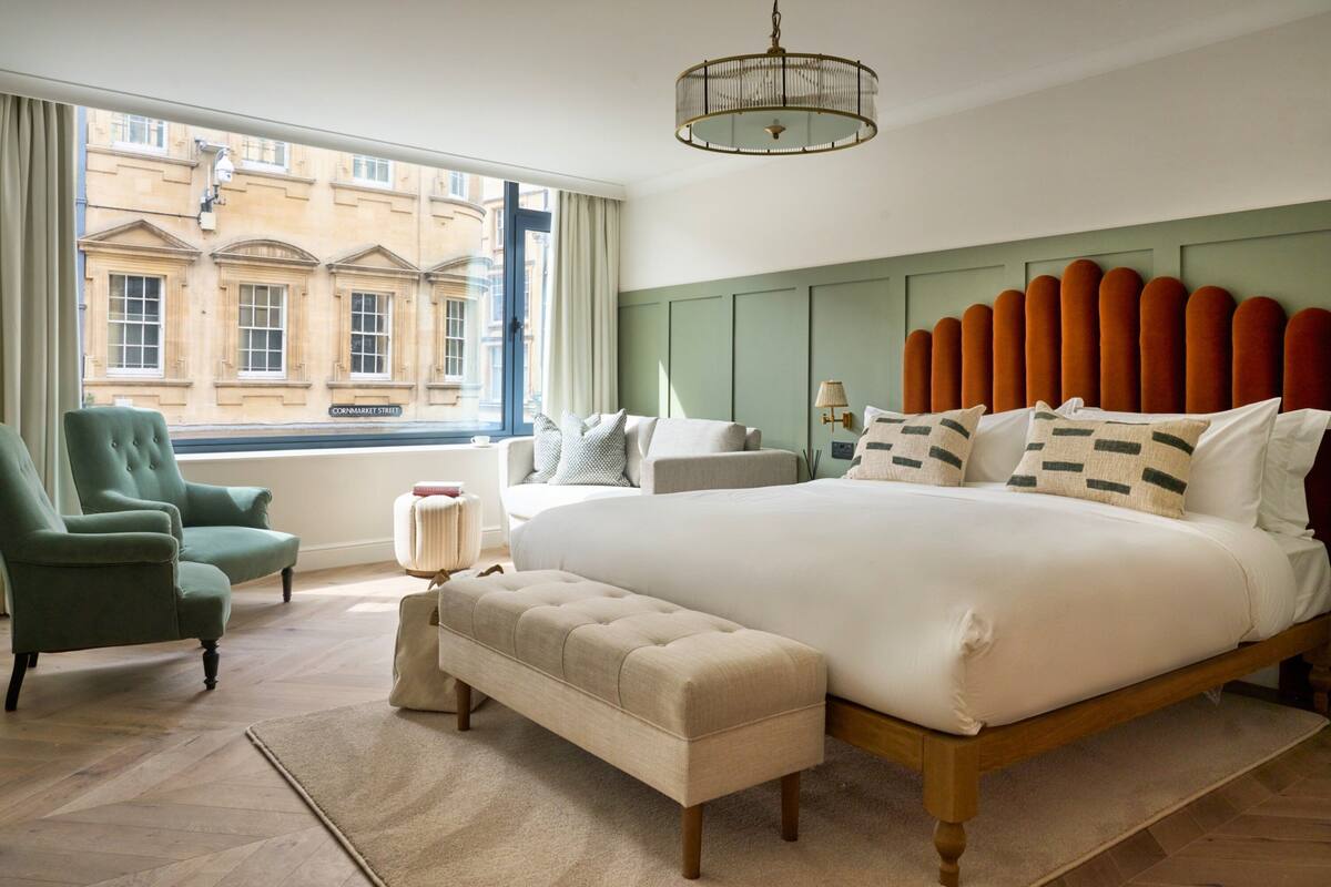 A bedroom with a large bed, a green armchair, a bench, and a view of a building through the window.
