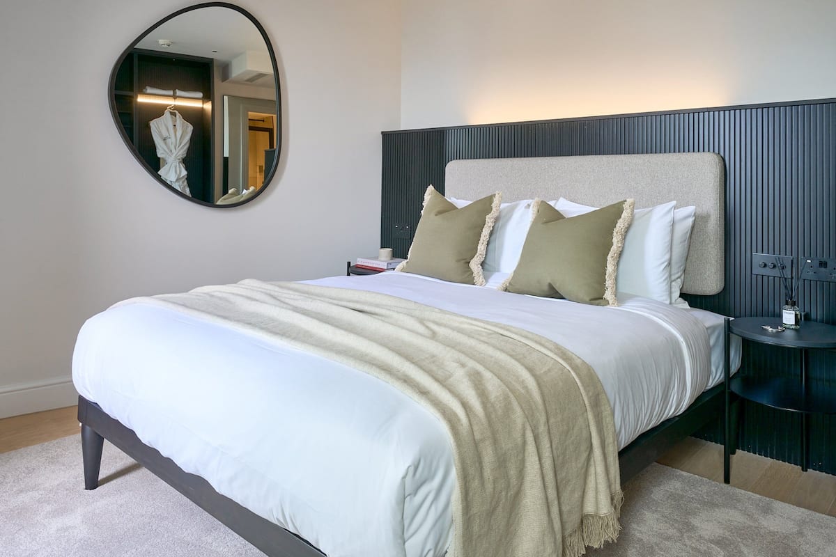 A neatly made bed with a white duvet, a beige throw blanket, and two pillows. A round mirror hangs on the wall above the bed.