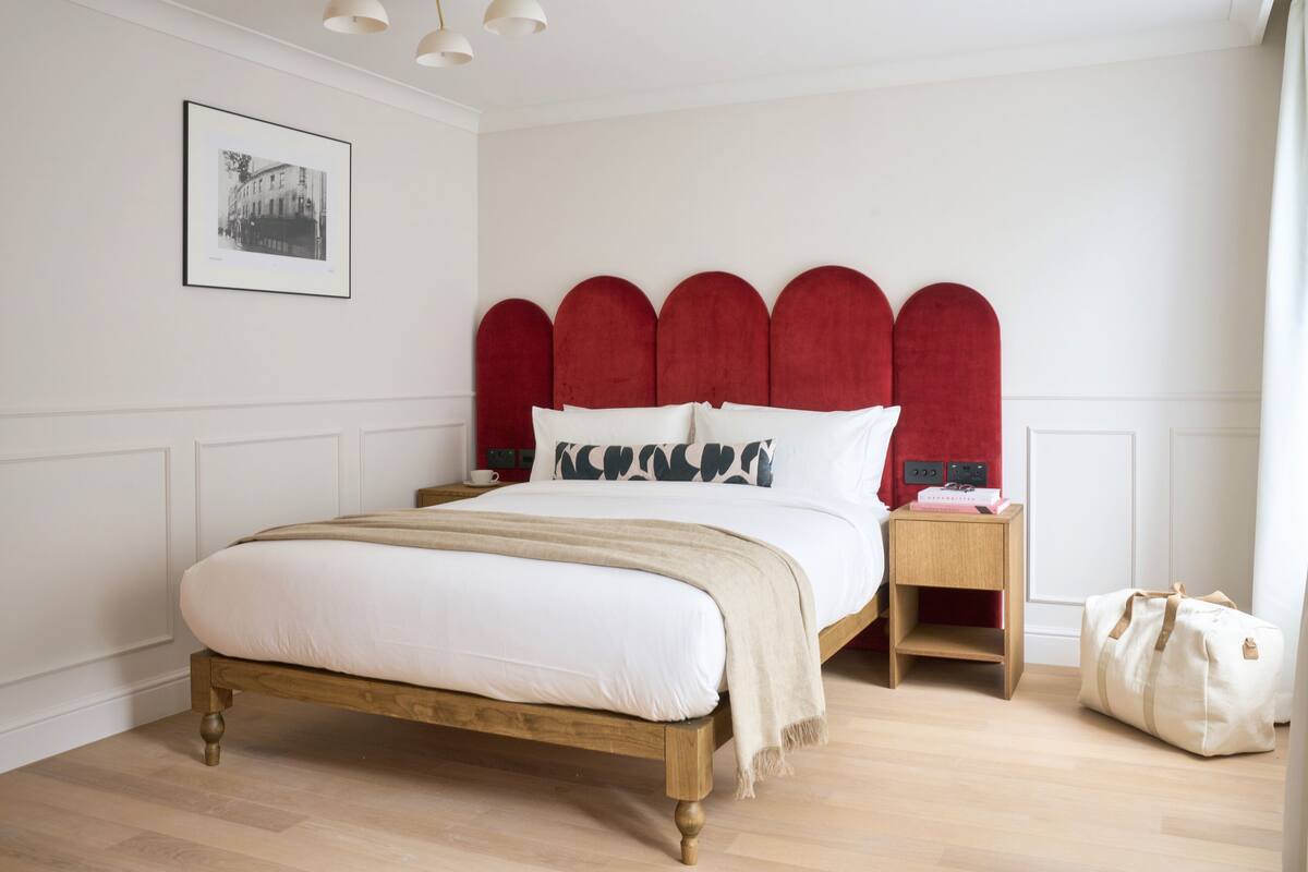 A bedroom with a wooden bed, red upholstered headboard, white bedding, a bedside table with a lamp, and a bag on the floor.