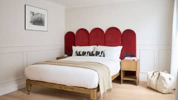 A bedroom with a wooden bed, red upholstered headboard, white bedding, a bedside table with a lamp, and a bag on the floor.