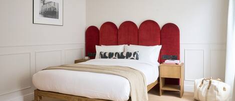 A bedroom with a wooden bed, red upholstered headboard, white bedding, a bedside table with a lamp, and a bag on the floor.