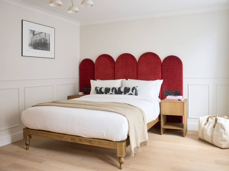 A bedroom with a wooden bed, red upholstered headboard, white bedding, a bedside table with a lamp, and a bag on the floor.